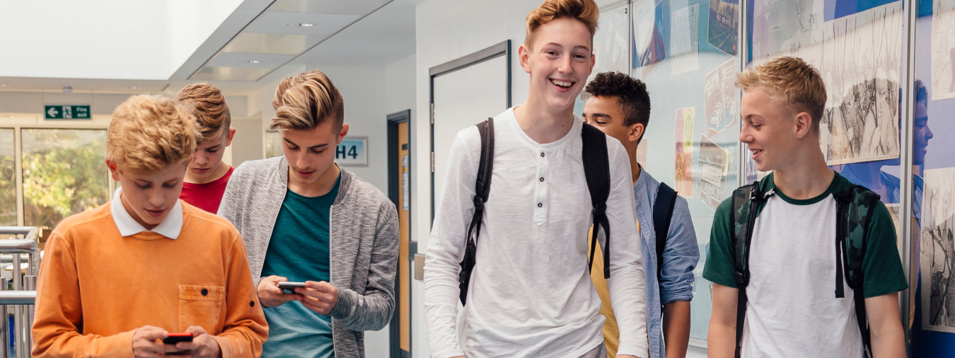 Eine Gruppe Jugendlicher geht durch einen Schulflur. Einige von ihnen schauen auf ihre Smartphones. Sie tragen Freizeitkleidung und Rucksäcke.