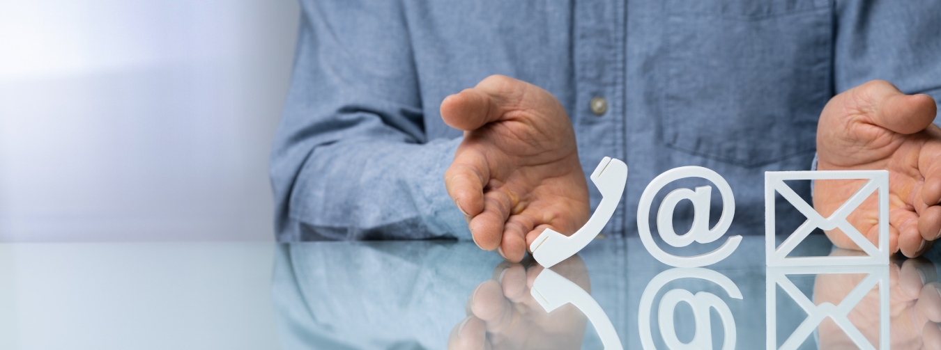 Eine Person in Hemd zeigt mit den Händen auf Symbole für Telefon, E-Mail und Briefumschlag auf einem Glastisch.