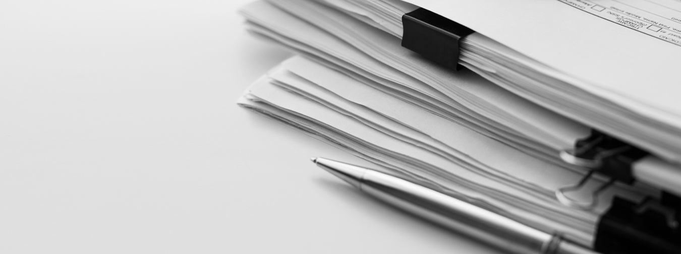 Ein Stapel Papierdokumente liegt auf einem Tisch. Daneben befindet sich ein Kugelschreiber. 